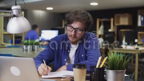 戴眼镜的年轻人正在办公室的笔记本上写东西视频的预览图