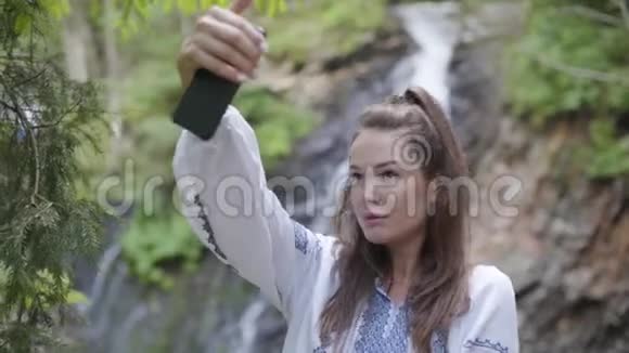 可爱的年轻女子穿着刺绣衬衫自拍站在瀑布前与大自然的联系视频的预览图