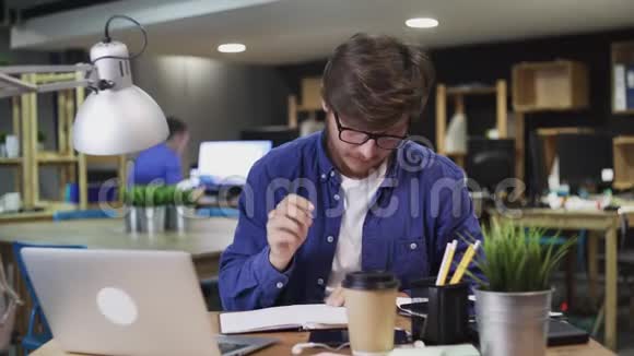年轻的商人在现代办公室工作记笔记输入笔记本电脑并打电话视频的预览图