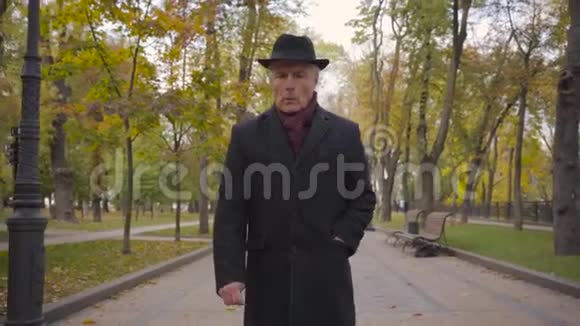 成熟的白种人一头白发戴着优雅的黑色帽子外套和勃艮第围巾漫步在小巷里视频的预览图