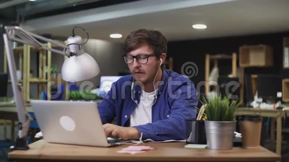 带耳机的年轻程序员在办公室听音乐在笔记本电脑上工作视频的预览图