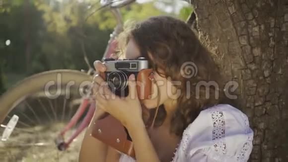 漂亮的年轻摄影师卷发坐在树下在花园或公园里用旧相机拍照视频的预览图
