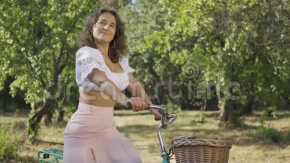 可爱的卷发年轻女子站在花园里或公园里骑着自行车看着镜头农村生活视频的预览图