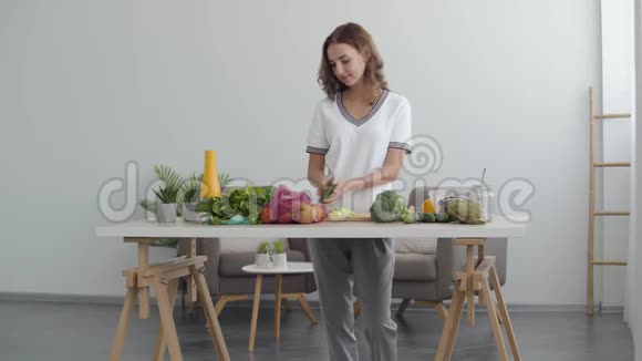 一个漂亮的女人在家中站在厨房的桌子旁用刀子在厨房的切割板上切黄瓜视频的预览图