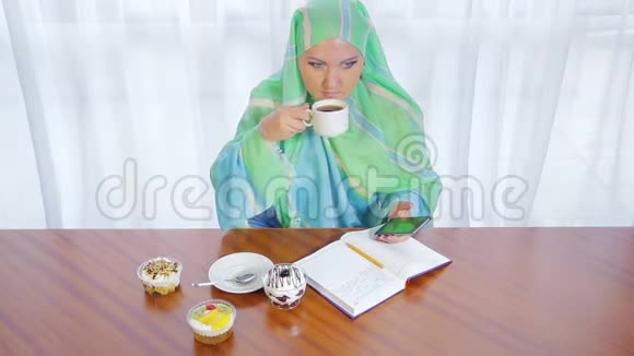 一个年轻的穆斯林妇女在一家咖啡馆里戴着一条浅色围巾用电话日记和喝茶远程工作视频的预览图