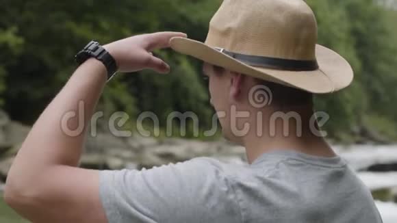 从后面看戴帽子的人看着站在森林里的河岸上在森林里徒步旅行的人视频的预览图
