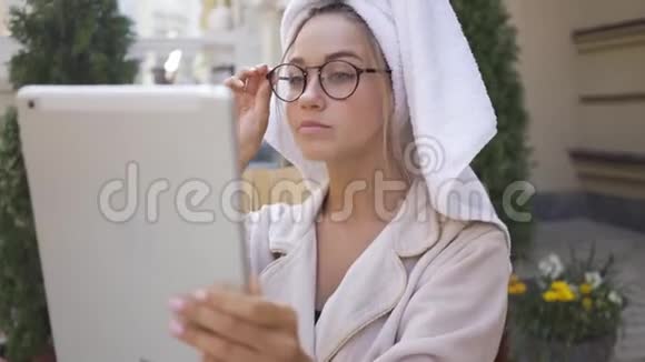 戴着眼镜穿着浴袍头上戴着毛巾坐在后院看着平板电脑的年轻女子的画像视频的预览图