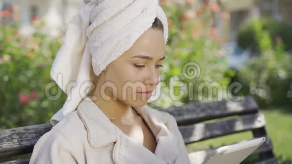 一位身穿浴袍头戴毛巾的年轻女子在公园里坐在长凳上时的肖像受到了震惊的消息视频的预览图