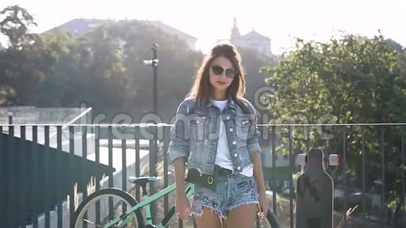 日落时骑自行车后穿着牛仔夹克时尚短裤和太阳镜在街上摆姿势的美丽黑发女孩视频的预览图