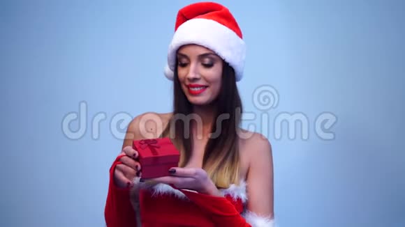 年轻漂亮性感的圣诞女孩穿着性感的红色连衣裙戴着可爱的圣诞帽打开一个红色小礼盒视频的预览图