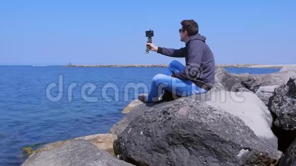 度假时男人坐在海边的巨石上用智能手机聊天视频的预览图