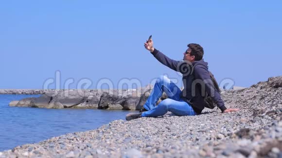 男人旅行者坐在海石海滩上在度假时制作自拍智能手机视频的预览图