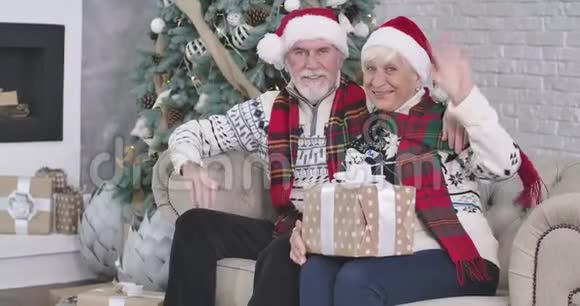 中镜头微笑的老白种人男人和女人坐在沙发上拿着礼物对着镜头挥手快乐圣诞老人视频的预览图