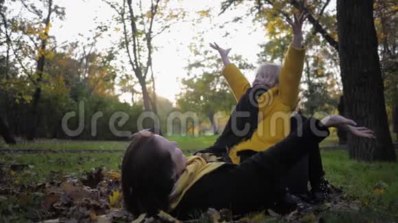 秋天快乐的妈妈和一个可爱的女儿躺在秋天公园的草坪上在花园散步时扔树叶视频的预览图