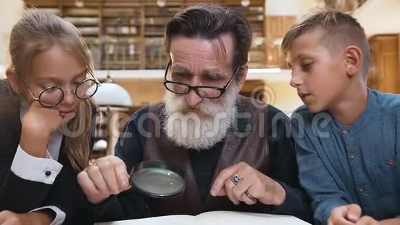 聪明的老胡子爷爷戴着眼镜带着他感兴趣的十几岁孩子用放大镜看书视频的预览图