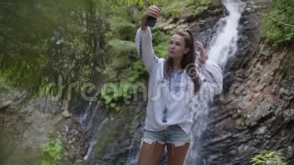 漂亮的年轻女人穿着刺绣衬衫自拍站在瀑布前面与大自然的联系视频的预览图