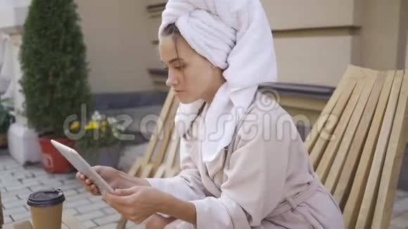 穿着浴袍头上有毛巾的漂亮年轻女人的肖像在后院用平板电脑坐在桌子旁自信视频的预览图