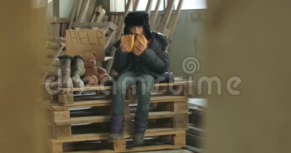 一个无家可归的女孩穿着脏衣服戴着帽子坐在调色板上吃着面包的画像视频的预览图