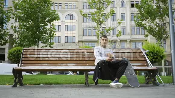 年轻人坐在公园的长凳上用手机听音乐拐杖和滑板就在附近视频的预览图