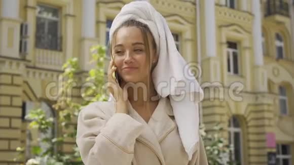 一幅可爱的微笑着的柔少女穿着浴袍头上戴着毛巾在街上用手机说话自信视频的预览图