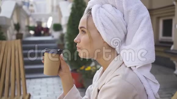 一幅年轻女子穿着浴袍头上戴着毛巾坐在后院喝咖啡的桌子旁自信的富人视频的预览图