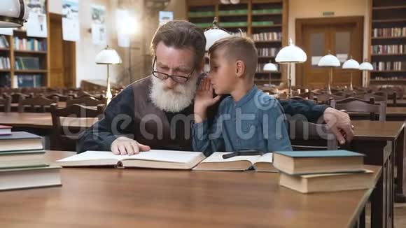 英俊的金发男孩对坐在图书馆里的有经验的老爷爷低语视频的预览图
