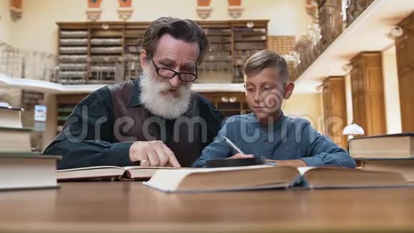 一位12岁的金发孙子和他那位年长的聪明的爷爷一起在图书馆看书的正面景色视频的预览图