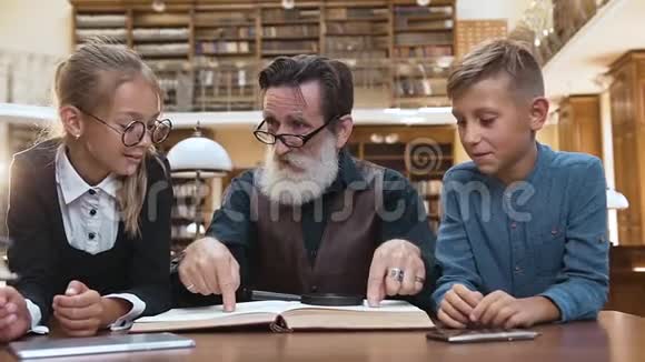 聪明的老人梳着整齐的胡须坐在图书馆的桌子旁和他可爱的少年一起看书视频的预览图