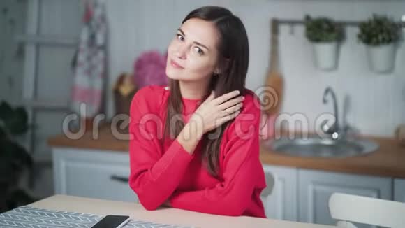 一幅可爱的年轻女子坐在家里的厨房里看着镜头微笑视频的预览图