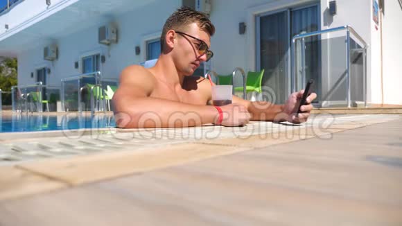 戴太阳镜的年轻人喝饮料在智能手机上拍照帅哥在酒店游泳池和视频的预览图