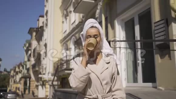 肖像可爱微笑的年轻女子穿着浴袍头上戴着毛巾走在街上喝着咖啡自信的女孩视频的预览图