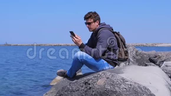 人类旅行者打电话给智能手机坐在石头上等待海上背景的回答视频的预览图