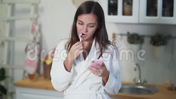 穿着白色浴袍的年轻女子在早晨刷牙使用智能手机视频的预览图