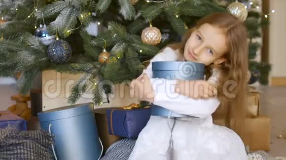 微笑着可爱的金发女孩拥抱着一个蓝色的礼物盒她坐在圣诞树旁快乐的童年和圣诞节视频的预览图