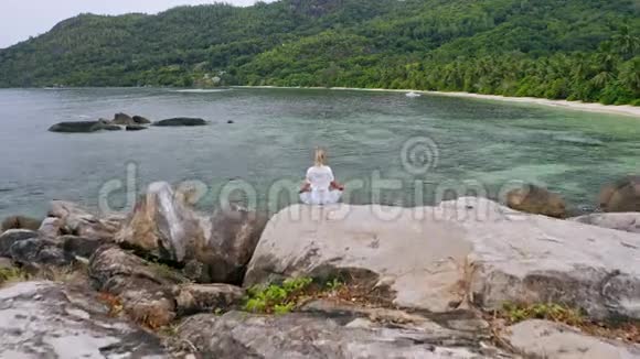 空中飞过一个金发晒黑的女孩坐在瑜伽姿势上在花岗岩岩石上冥想对抗异国情调的热带泻湖视频的预览图