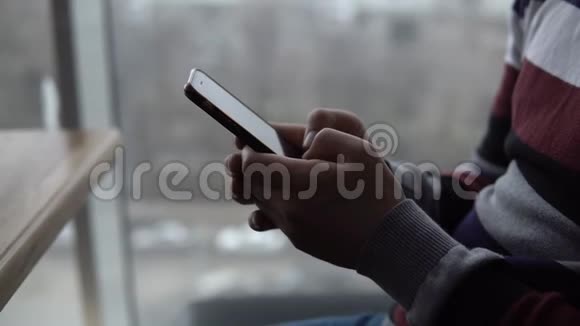 一个有手机特写的年轻人一个男人坐在全景窗旁的咖啡馆里手里拿着一部智能手机从这里看视频的预览图