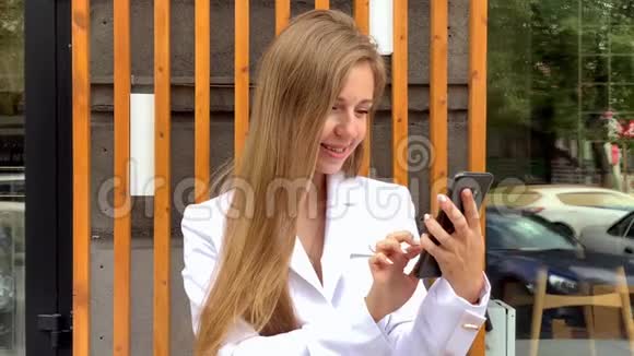 穿着白色夹克的快乐年轻美丽微笑的女人在户外使用智能手机视频的预览图