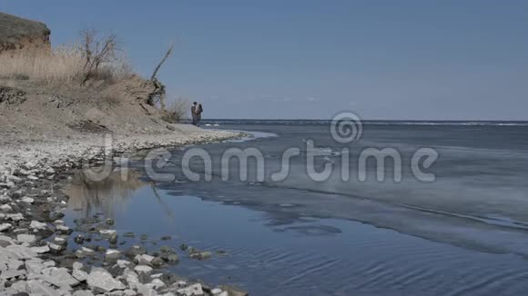 一对年轻夫妇沿着冰封的河流的岸边散步阳光明媚的春天视频的预览图