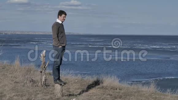一个年轻人站在树桩上在一条河的背景下思考着阳光明媚的日子视频的预览图