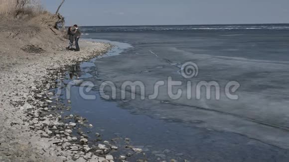 一对年轻夫妇沿着冰封的河流的岸边散步阳光明媚的春天视频的预览图