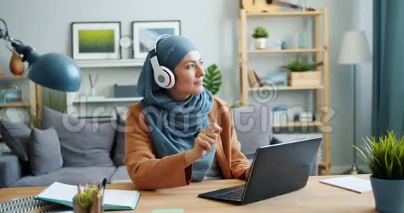 戴着头巾的混血女孩在家里用笔记本电脑在耳机里享受音乐视频的预览图
