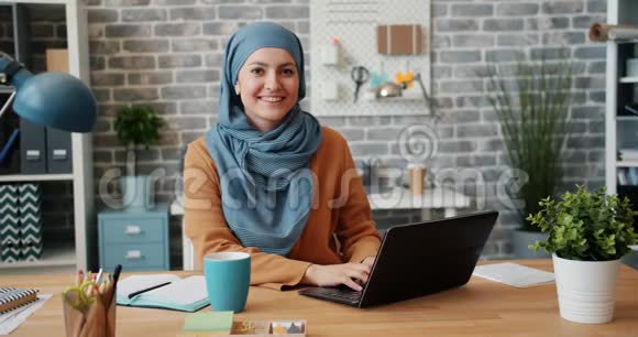 戴头巾的穆斯林女孩微笑着在办公桌前看镜头的画像视频的预览图