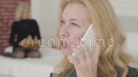 一位迷人的白种人成熟女性的特写肖像在电话旁交谈微笑她的金发朋友坐着视频的预览图