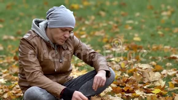 迷人的家伙坐在公园里的落叶上仔细地拿起一片黄色的枫叶仔细地检查它视频的预览图