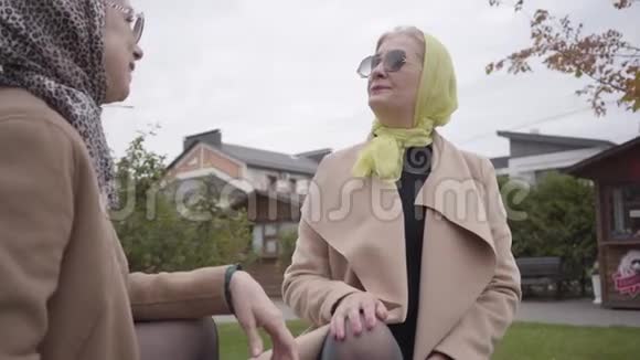 两名戴着头巾和太阳镜的白种人成年女性坐在长凳上聊天优雅优雅视频的预览图