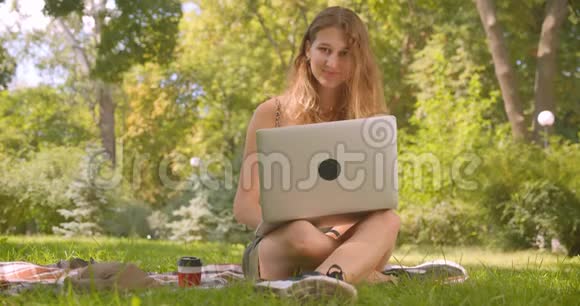 年轻漂亮的白种人女性使用笔记本电脑开心地笑着看着坐在草地上的摄像机视频的预览图