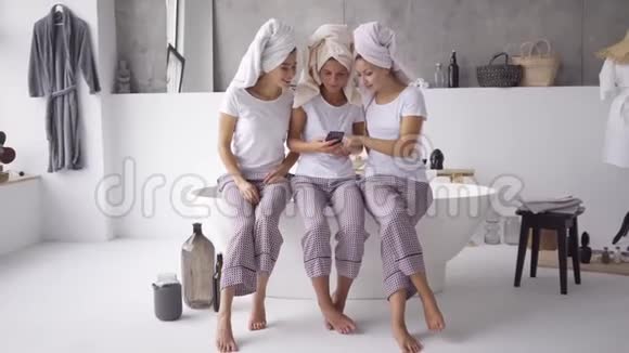 三个穿着相同睡衣和毛巾的可爱女朋友一起在家看手机视频的预览图