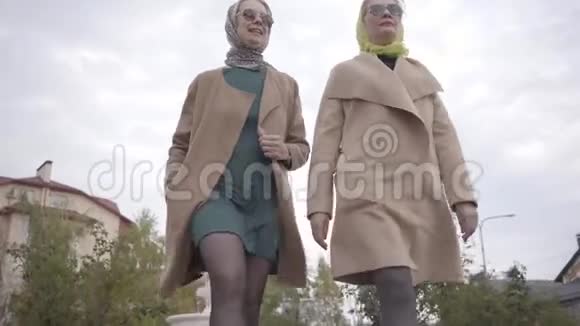 两名身穿米色外套头戴头巾和太阳镜的成年白人妇女在城市公园散步的俯视图视频的预览图