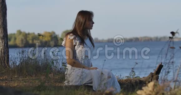 神秘的高加索女人坐在湖岸上环顾四周穿着浅色白裙休息的漂亮女孩视频的预览图