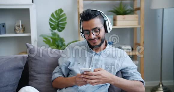 英俊的阿拉伯男人在家里用智能手机耳机听音乐视频的预览图